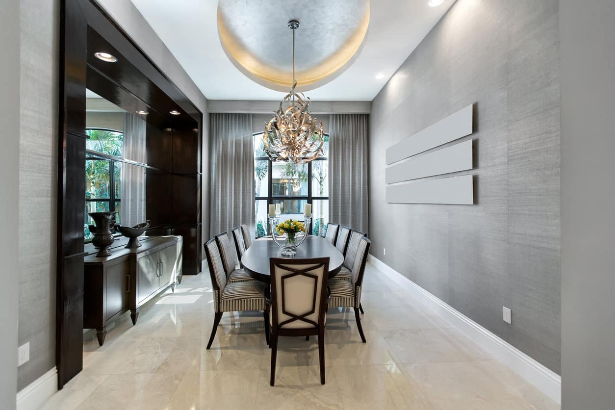 An eccentric chandelier that hung from the round tray ceiling illuminates this dining room featuring an oval dining table and stripe cushioned chairs over marble tile flooring. It is accompanied by a wooden buffet table with antique vases on top.
