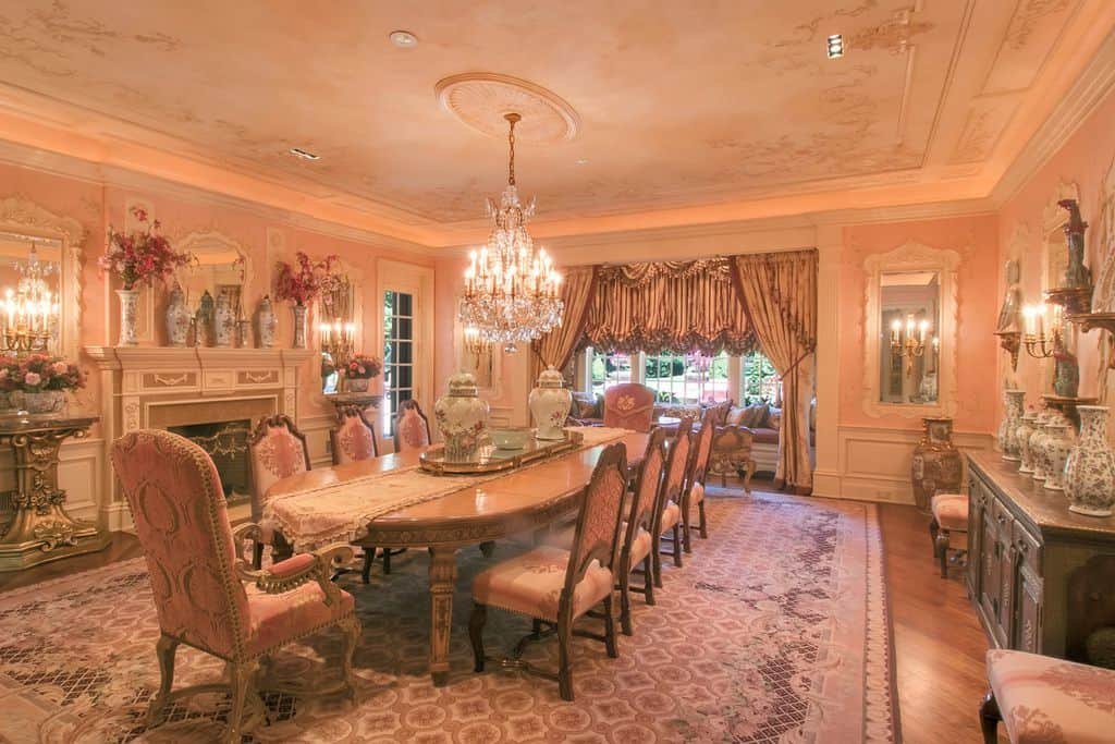 Charming dining room filled with ornate tables and elegant seats along with a glamorous crystal chandelier that hung from the tray ceiling. It is decorated with ceramic vases and lovely mirrors mounted above the white wainscoting.